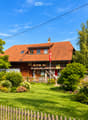Two-story house with red-tiled roof, wooden balcony, and well-maintained garden with a wooden fence.