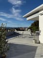 Modern rooftop terrace with tiled floor, small table and chairs, potted plants, and a view of the city and mountains.