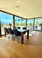 Bright dining area with wooden floor, large windows offering a scenic mountain view, dining table with chairs, decorative shelf, and potted plants.