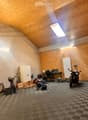 A spacious workshop or garage with a slanted wooden ceiling, exposed brick wall, and checkered floor. Contains a desk with a chair, a motorcycle, a bicycle, a fan, a radiator, and various storage items.
