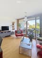 Modern living room with wooden flooring, large windows offering a view, a sofa, armchair, coffee table, dining table, TV, bookshelf, and potted plants.