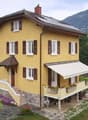 Two-story yellow house with white-framed windows, brown shutters, and a balcony with flower boxes. Solar panels on the roof and a scenic mountain view in the background.