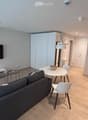 Modern living room with a grey sofa, coffee table, dining table, chairs, and a television mounted on the wall. The room features light wooden flooring and white cabinets.