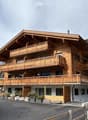 A large, multi-story wooden house with multiple balconies, situated in a scenic mountainous area with a paved driveway and surrounding greenery.