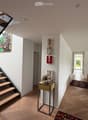 Modern hallway with wooden flooring, a staircase leading upwards, and artwork on the walls. A small table with a floral arrangement is placed near the staircase, and a living area with a sofa is visible at the end of the hallway.