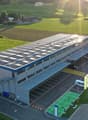 A modern industrial building with a flat roof, solar panels, and a parking lot, surrounded by green fields and agricultural structures.