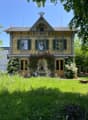 Two-story house with yellow and grey exterior, decorative wooden trim, multiple windows, and a well-maintained garden with outdoor furniture.