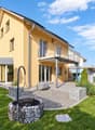 Two-story yellow house with a modern design, featuring a balcony, large windows, and a well-maintained garden with a stone fire pit.
