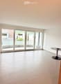 Empty room with white tiled flooring, large sliding glass doors leading to a balcony, and a small round table with books in the corner.