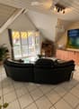 Bright living room with a black sectional sofa, coffee table, wooden cabinet, and a fireplace. Large windows offer a view of the garden and pool.