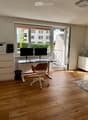 Bright, modern room with wooden flooring, large windows, and a balcony. Contains a desk with two monitors, a chair, a small table with chairs, a printer, and a fan.