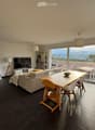 Modern living and dining area with a sofa, dining table, chairs, television, shelves, and a large window offering a scenic view.