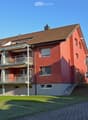 red building with several balconies, gray walls, brown roof, brick ground, garden in front