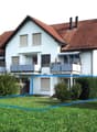 Two-story house with red-tiled roof, white exterior, multiple windows, balconies, and surrounding greenery.