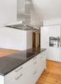 Modern kitchen with white cabinets, granite countertops, induction cooktop, built-in oven, and range hood. Wooden flooring and large window with view of greenery.