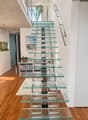 Modern hallway with a sleek glass staircase featuring metal railings, wooden flooring, and contemporary decor including a small shelf unit and artwork.