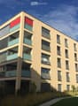 Modern multi-story residential building with light beige facade, large windows, and balconies. Features a raised ground floor and a sign reading "A VENDRE" (For Sale).