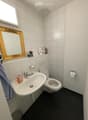 A small bathroom with a white sink, toilet, and mirror. The sink has a silver faucet and a soap dispenser. Toilet paper and a towel are visible, along with a decorative mirror and a small figurine on the windowsill.