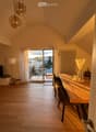 Bright, airy room with wooden flooring, large window leading to a balcony, dining table with chairs, decorative vase, and a TV on a stand.