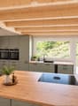 Modern kitchen with grey cabinets, wooden island, induction cooktop, double oven, and large windows overlooking a garden.