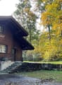 A two-story wooden house with a stone wall and steps leading to the entrance, surrounded by autumn foliage and trees.