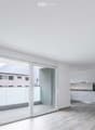 Bright, empty room with large sliding glass doors leading to a balcony, white walls, light wooden flooring, and a small adjacent kitchen area with white cabinets and appliances.