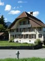 Two-story house with a brown tiled roof, multiple windows with shutters, and a satellite dish on the roof. The house is surrounded by a low stone wall and a grassy area with trees and mountains in the background.