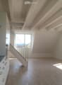 Open-plan space with a modern kitchen area on the left, a staircase leading upwards, and large windows allowing natural light to flood the room. The floor is light-colored wood, and the walls are white.