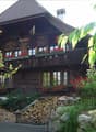 Traditional wooden house with a steep roof, multiple windows, and flower boxes on the balconies. The house is surrounded by greenery and a stack of firewood is visible in front.