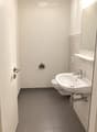 Modern bathroom with a white sink, chrome faucet, toilet, and a mirror above the sink. The walls are tiled in light beige, and the floor is covered with dark gray tiles.