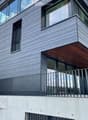 Modern multi-story building with grey brick facade, large windows, and a wooden balcony with metal railing.