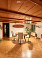 A modern dining area with a round wooden table, four chairs, potted plants, a shelf with decorative items, and a surfboard on the wall.