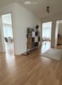 Bright hallway with wooden flooring, leading to multiple rooms including a kitchen, dining area, and living room. Features a small shelf with decorative items and a rug in the center.