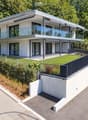 Modern two-story house with white exterior, large glass windows, and a raised ground floor. Features include a balcony, garage, and landscaped garden area.