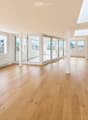 Bright, empty room with light wooden flooring, large windows, and sliding glass doors leading to a balcony. The space features white walls, a skylight, and a radiator on one wall.