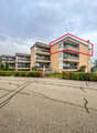 Multi-story residential building with balconies and modern design, surrounded by greenery and a paved parking area.