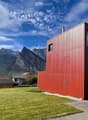 Modern red metal house with large windows, surrounded by a green lawn and mountains in the background.