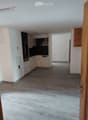 Empty room with light wooden flooring, white walls, and a small kitchen area with cabinets and appliances visible in the background.