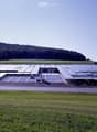 Large industrial building with solar panels on the roof, surrounded by open fields and a parking area.