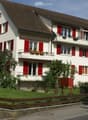 A two-story house with white walls, red shutters, and a brown roof. The house features multiple windows, balconies, and is surrounded by greenery and a wooden fence.