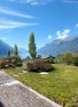 A serene garden with a wooden deck, stone bench, lush greenery, and a scenic mountain and lake view in the background.