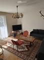Living room with a dark grey sofa, coffee table, red chairs, a chandelier, and a television on a stand. The room features a patterned rug, wooden flooring, and large windows with sheer curtains.