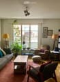 A cozy living room with a grey sofa, wooden coffee table, armchair, bookshelf, and decorative plants. The room features large windows with a view, warm lighting, and a mix of modern and classic furniture.