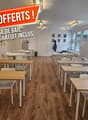 Bright, modern room with wooden flooring, multiple tables and chairs arranged for a collaborative workspace, shelves, a refrigerator, a printer, and a whiteboard.