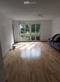 Bright, empty living room with wooden flooring, large windows, and a sofa with some items on it.