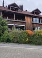 Two-story wooden house with a tiled roof, multiple balconies, and surrounding greenery.