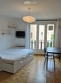 Cozy studio with a bed, small table, chairs, sofa, TV, and a refrigerator. Features wooden flooring and large windows leading to a balcony.