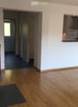 Bright hallway with wooden flooring, white walls, and a modern wood-burning stove on the left. The hallway leads to a kitchen area visible on the right with white cabinets and a countertop.