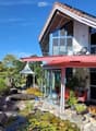 Modern two-story house with a red roof, large windows, and a spacious garden area featuring a pond, plants, and outdoor furniture.