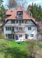 Two-story house with red-tiled roof, white walls, multiple windows, balconies, and a small garden area with outdoor furniture.